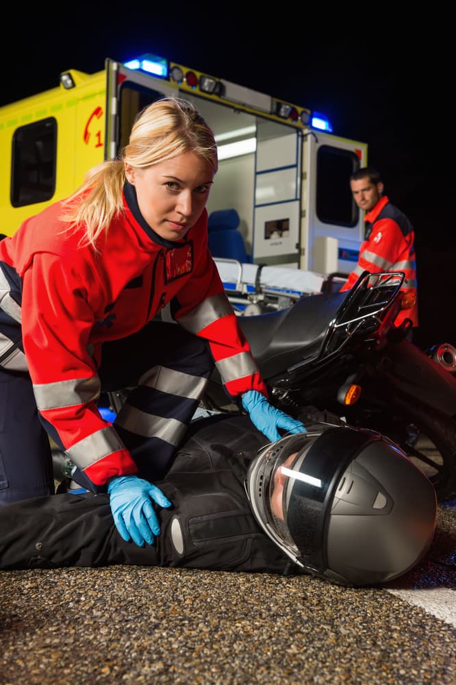 A paramedic assisting an injured motorcyclist at night with an ambulance in the background, representing emergency response after a motorcycle accident
