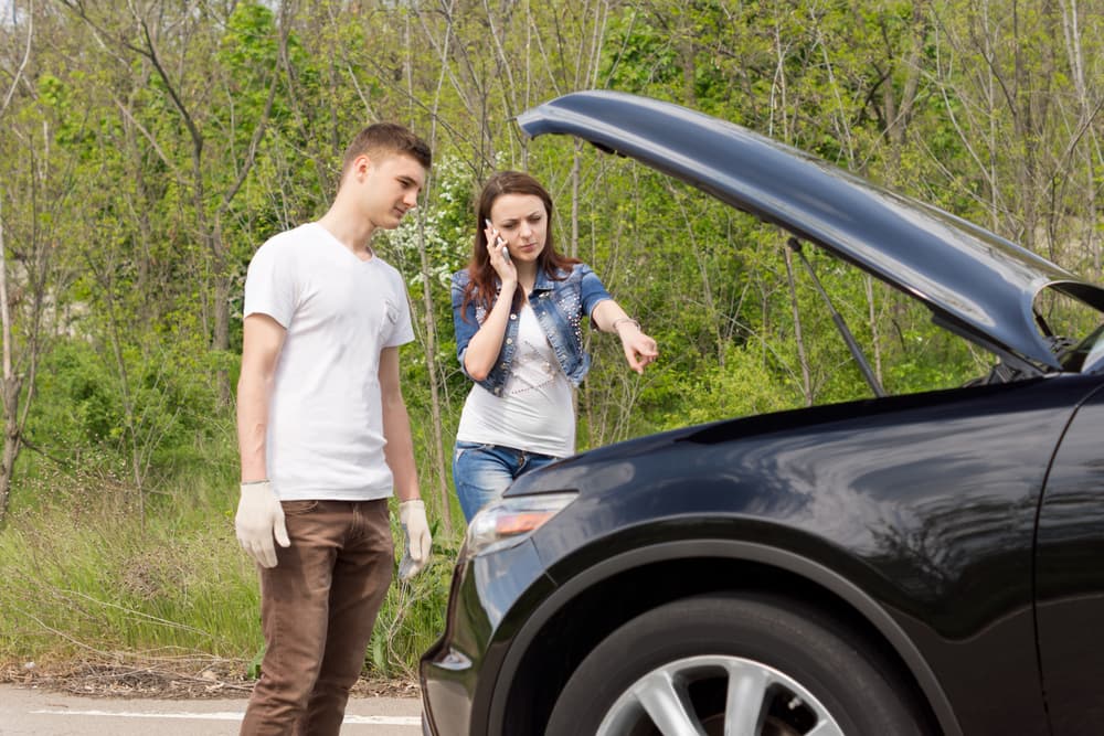 Young couple stranded on roadside, hood up, calling for help and pointing at car engine. Vehicle breakdown assistance in progress.