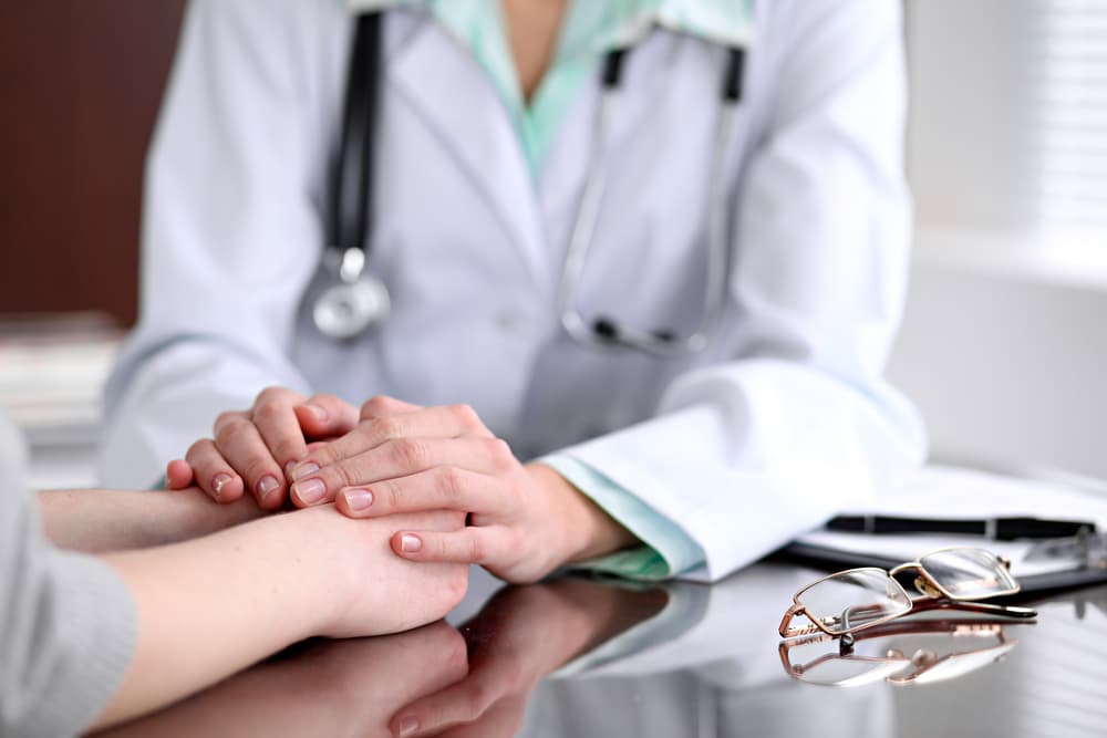 Doctor in a lab coat comforts patient by holding their hands on a desk, glasses nearby.
